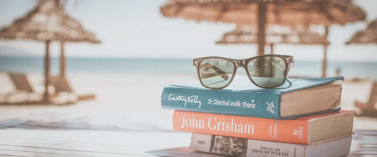 A small stack of books on the beach front, with a pair of reading glasses on top